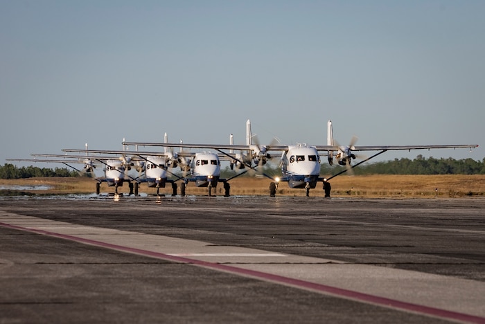 Four aircraft roll across cement in a straight line in close proximity to one another with a grassy backdrop