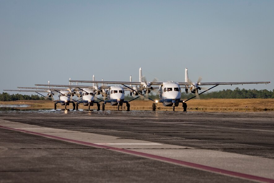 Four aircraft roll across cement in a straight line in close proximity to one another with a grassy backdrop