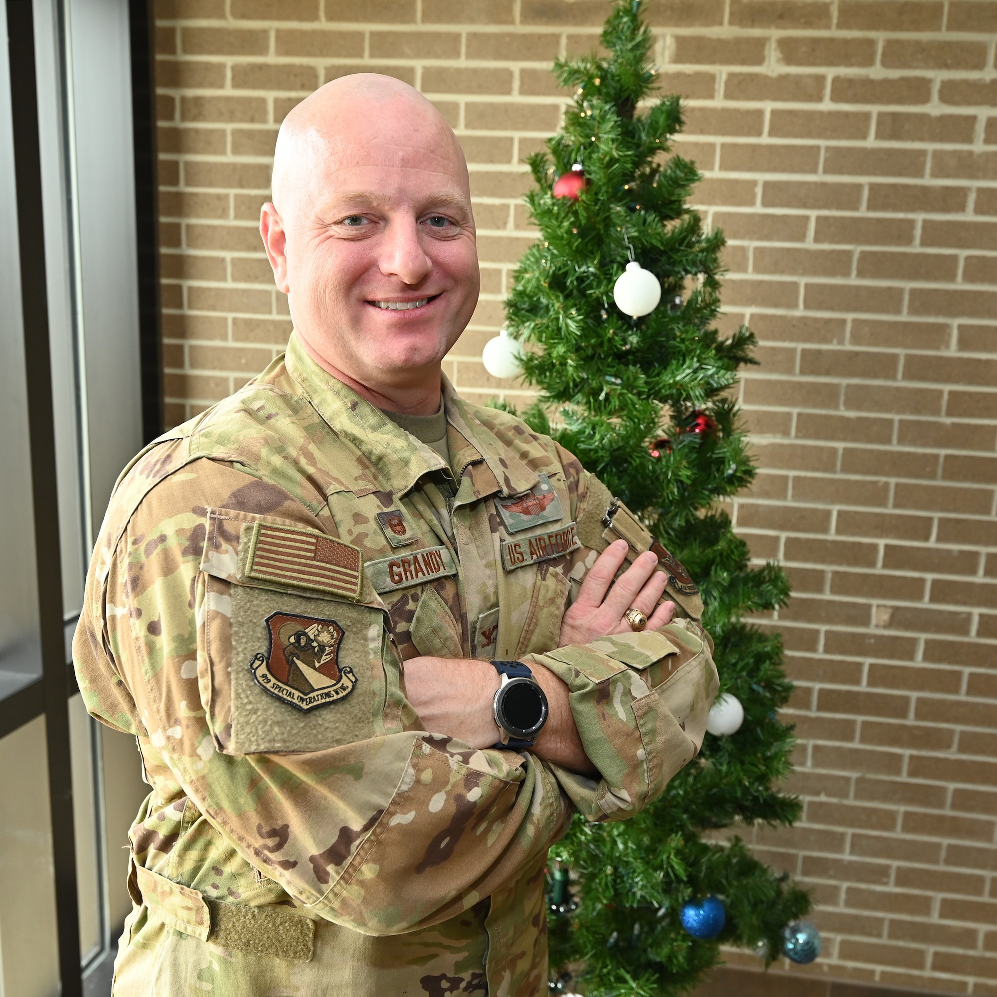 Airman stands in front of Christmas tree with brick wall background.