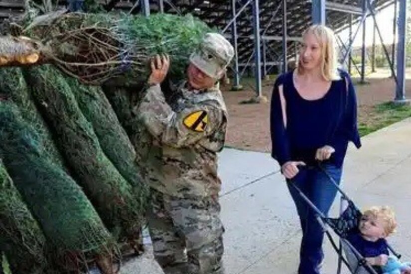 A service member has a Christmas tree on his shoulder, a woman with a baby in a stroller stand next to him.