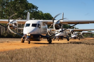 Three smaller aircraft with spinning propellers are parked in a straight line along a small dirt taxiway with grass in the foreground and trees in the background. A slight orange tint is reflected by the bottom of the airfoil from the dirt.