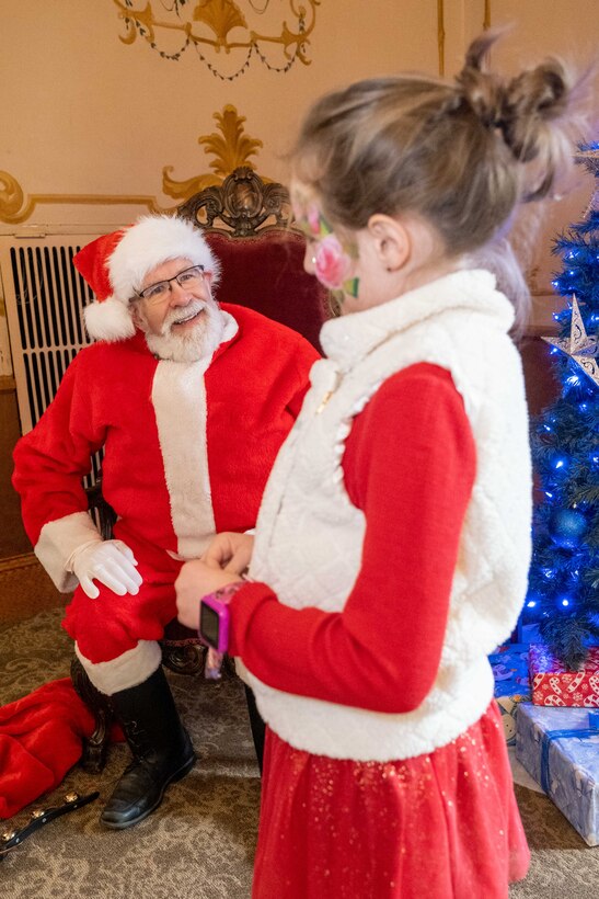 Santa sitting in a chair talking to a young girl.