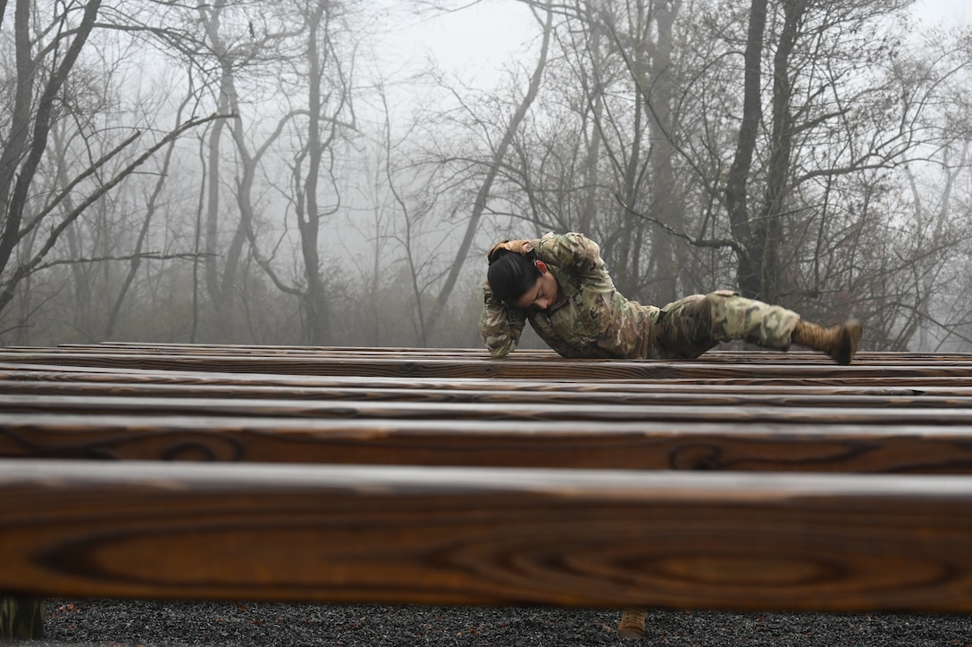 A soldier with her hands over her head climbs over a wooden bar.