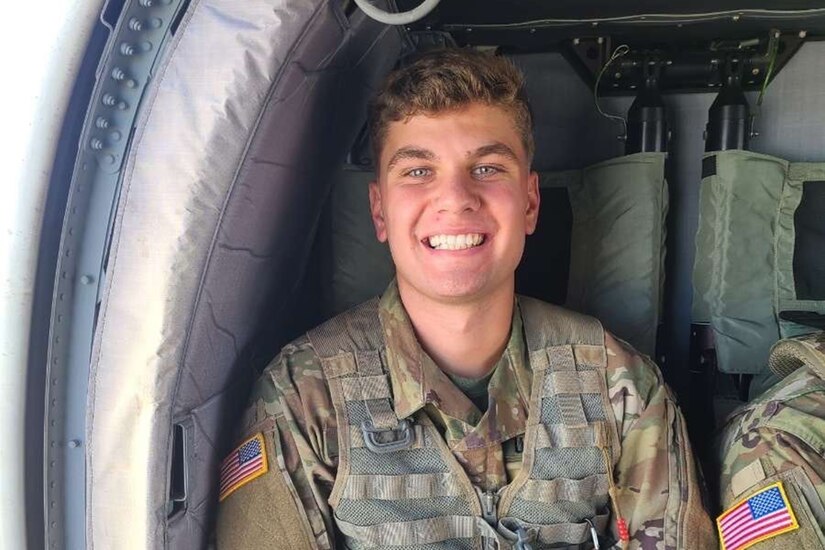 A soldier is shown from the chest up, seated in an aircraft, smiling.