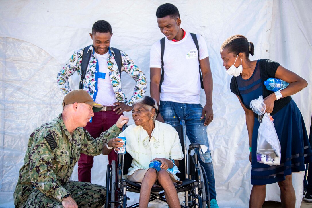 A sailor kneels to speak to a patient sitting in a wheelchair.