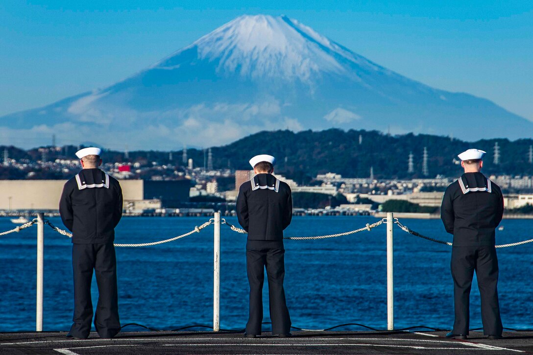 Three sailors stand around the rails of a ship; a mountain can be seen in the background.