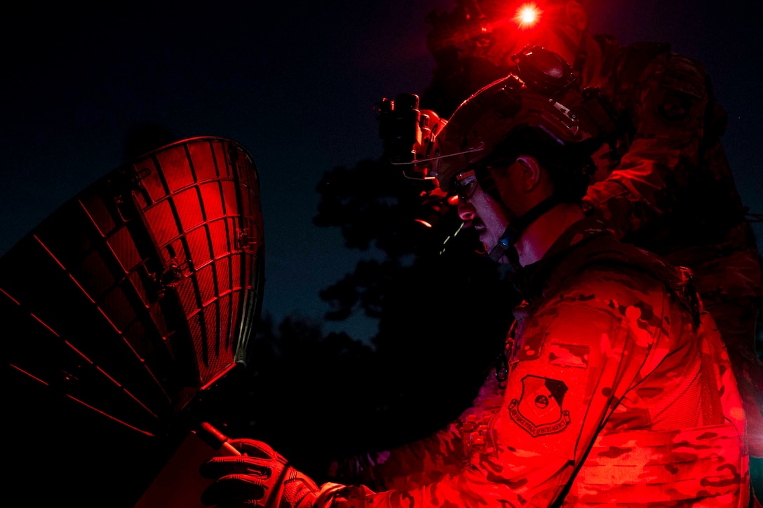 An airman illuminated by red light assembles a satellite.