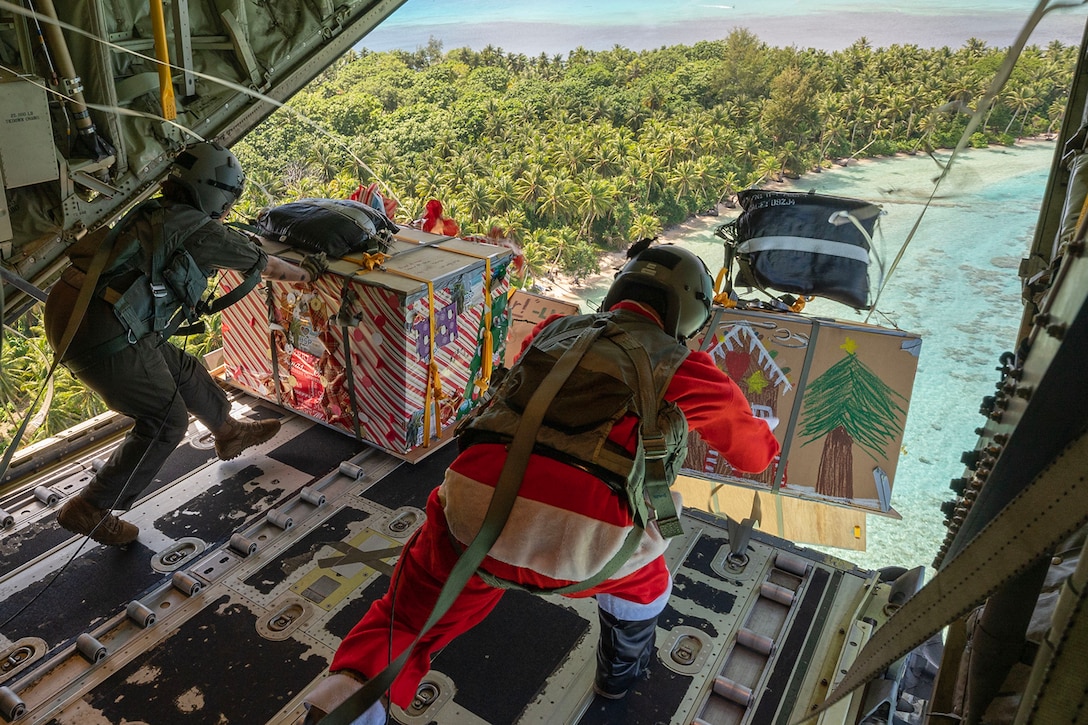 Airmen, one dressed as Santa, push decorated cardboard boxes out of an open aircraft.