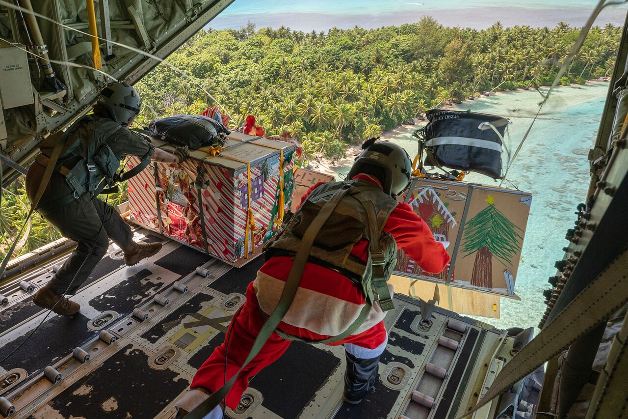 Airmen, one dressed as Santa, push decorated cardboard boxes out of an open aircraft.