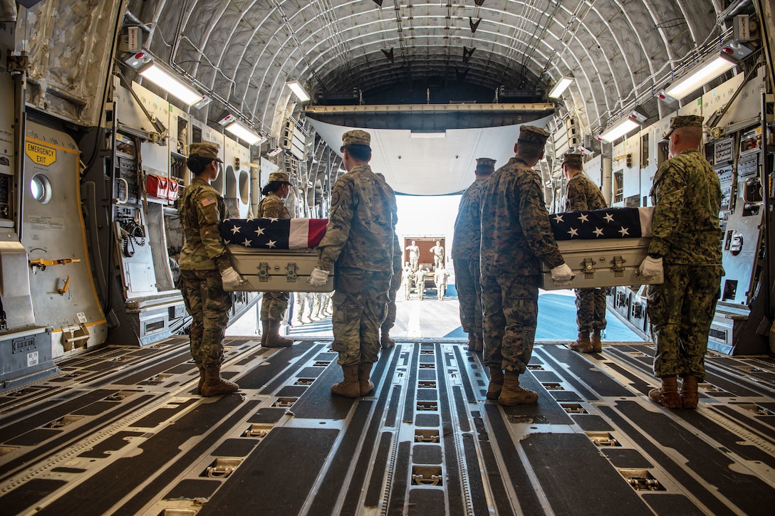 Service members carry transfer cases covered by U.S. flags out of a large aircraft.