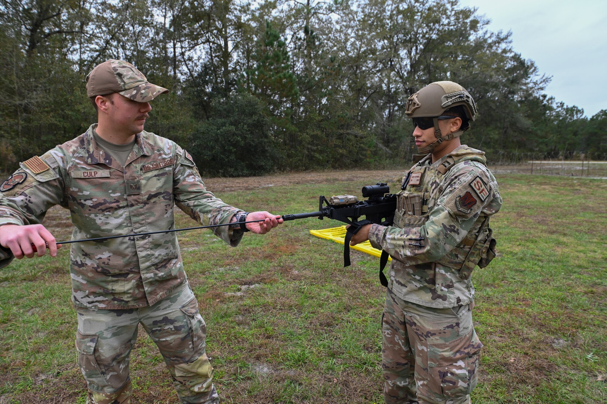 A photo of two Airmen in a field. The one on the left is using a long metal rod to stick into the opening of the M-4 that the other Airman is holding.
