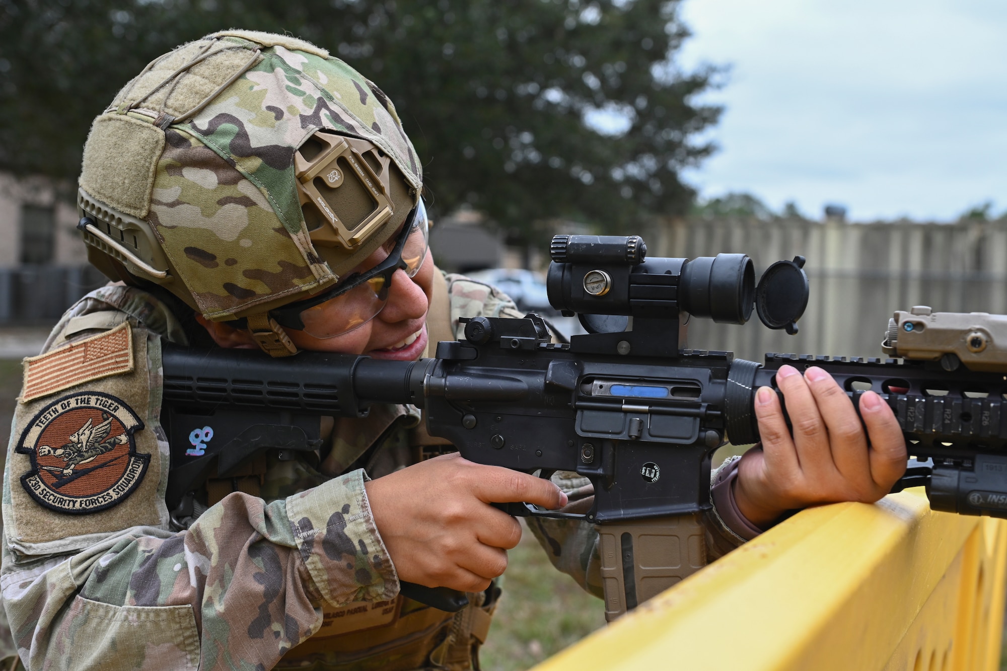 A photo of a close up of a woman holding an M-4 Carbine, resting her hand and weapon on top of a yellow barrier aiming towards the right side of the photo.