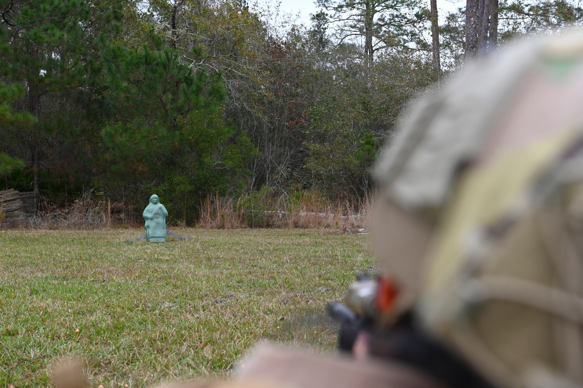A photo of a green human-like training dummy in a field with an Airman pointing an unloaded M4 Carbine at it.