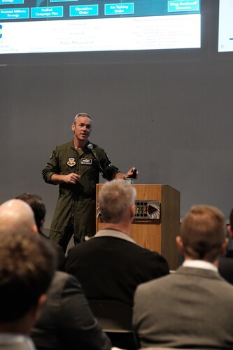 a military member in uniform stands next to a podium while civilians sit and listen