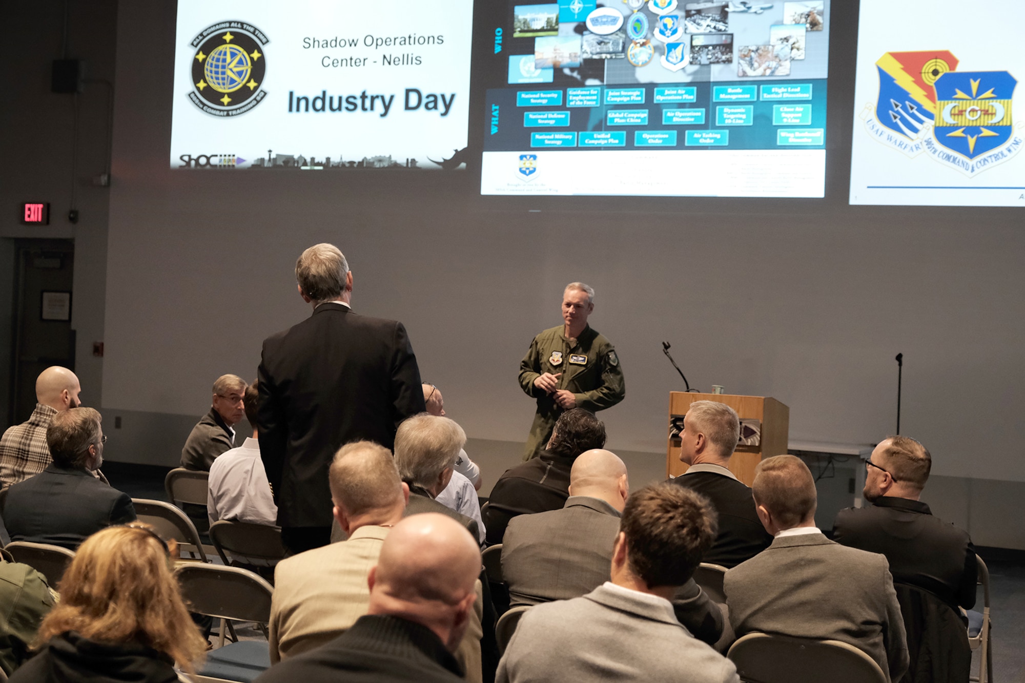 a military member in uniform stands next to a podium; on civilian stands asking a question while a large group of civilians sit and listen while threeslides are projected on a large wall