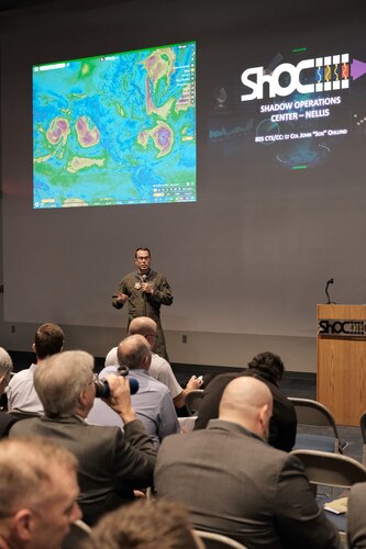 a military member in uniform stands next to a podium while a large group of civilians sit and listen while two slides are projected on a large wall