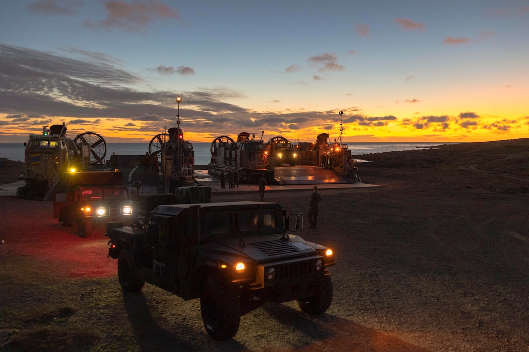 Marine Corps vehicles exit a small boat under a sunlit sky.