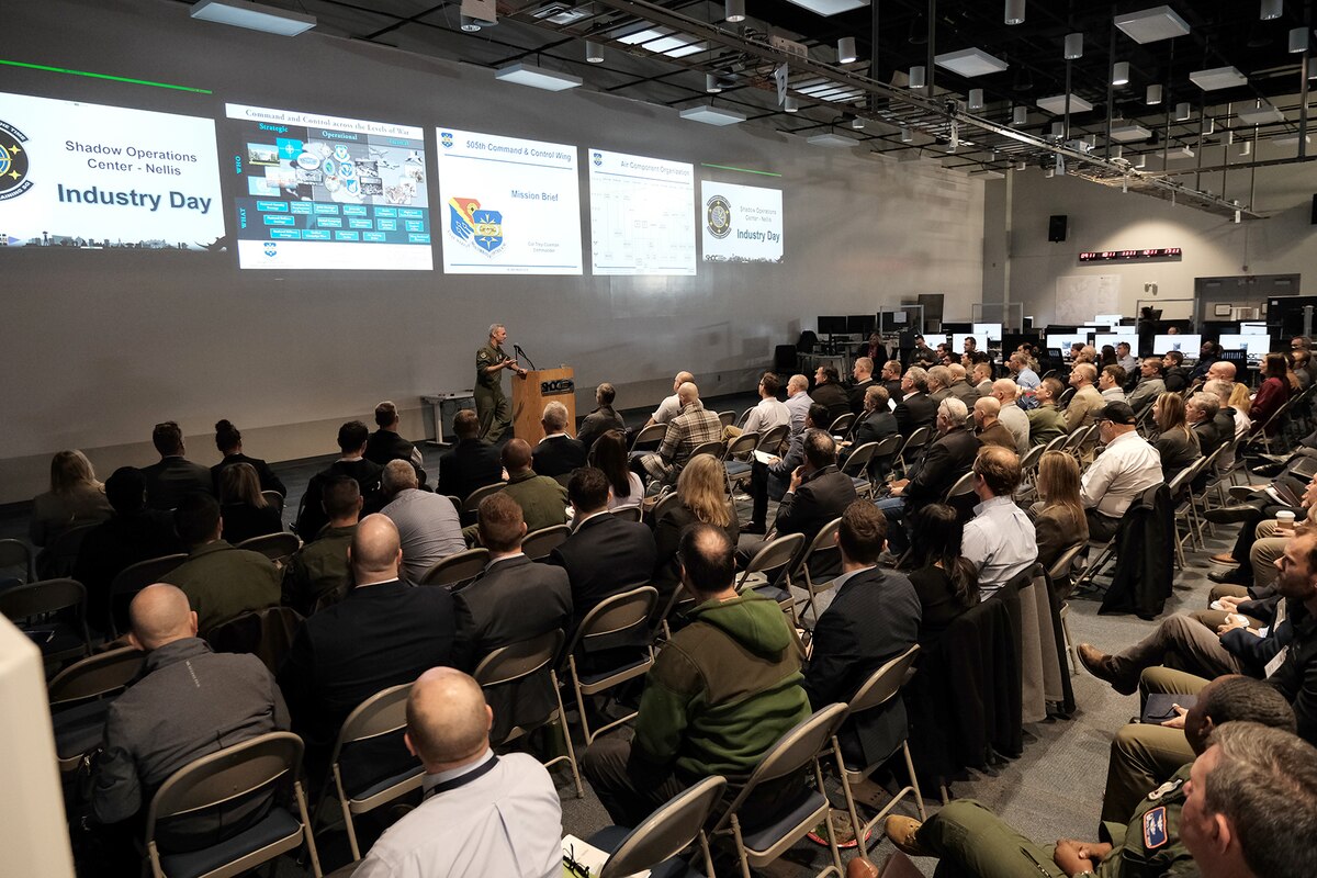 a large group of civilians and military sit in chairs listening to a military speaker with five slides displayed on the wall