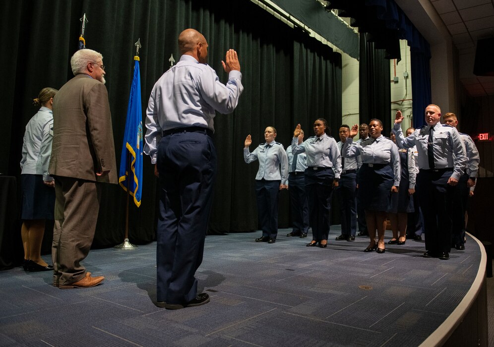 A man, with his back to the camera and his right arm raised, stands in front of a group of people with their arms raised. All of them are in Space Force uniform.