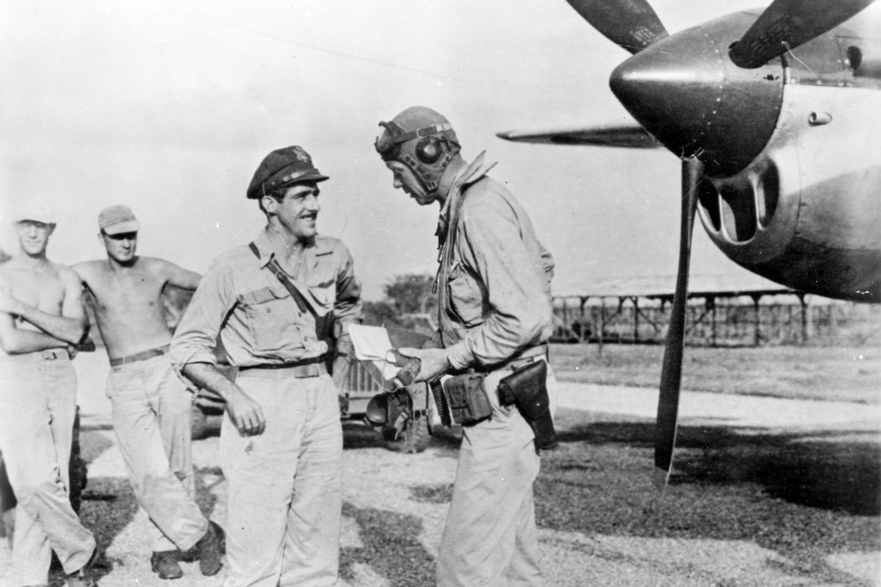 Two men talk near a propeller airplane while two other men lounge in the background.