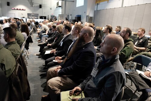 a large group of civilians and military sit in chairs listening to a military speaker