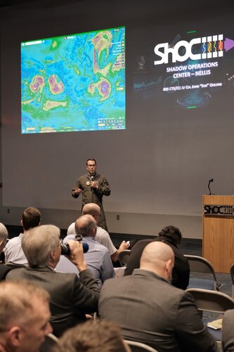 a military member in uniform stands next to a podium while a large group of civilians sit and listen while two slides are projected on a large wall