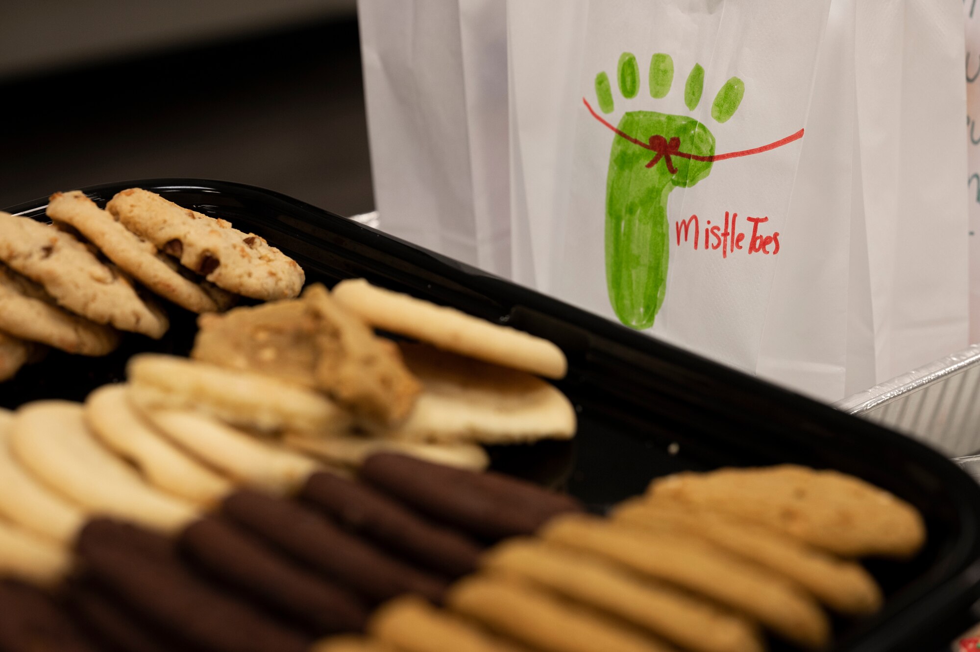 A tray of assorted cookies sits in the foreground of the photo with a white paper bag behind it.