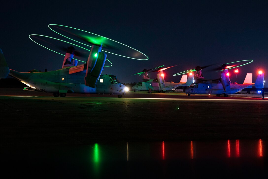 Three Marine Corps aircraft sit on a tarmac in the dark illuminated by colorful lights.