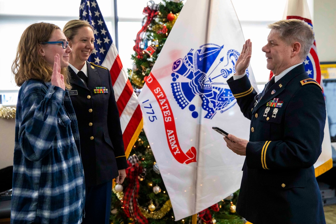 A woman holds her right hand up in front of a soldier holding up his right hand up as another soldier watches.