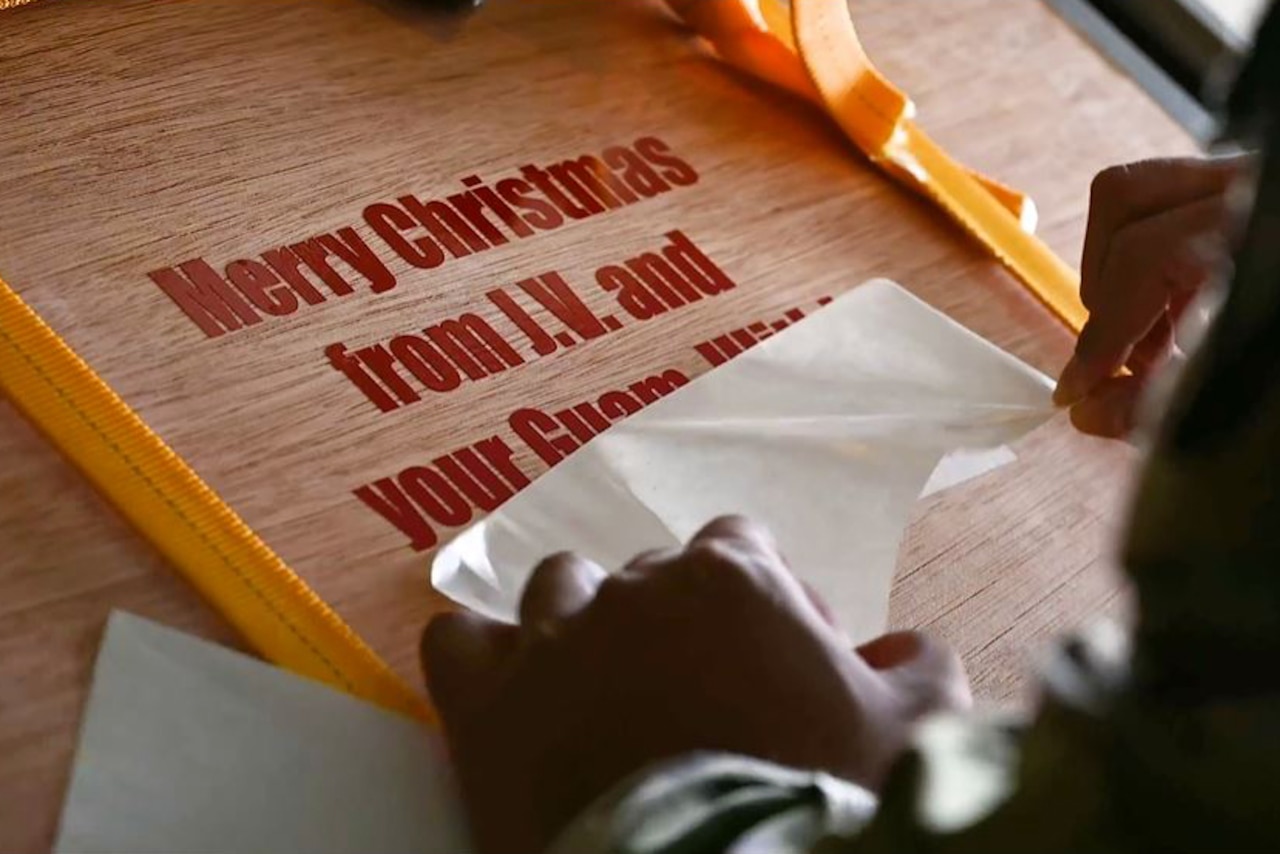 A service member peels back a label on a box that reads "Merry Christmas from J.V. and your Guam ..."