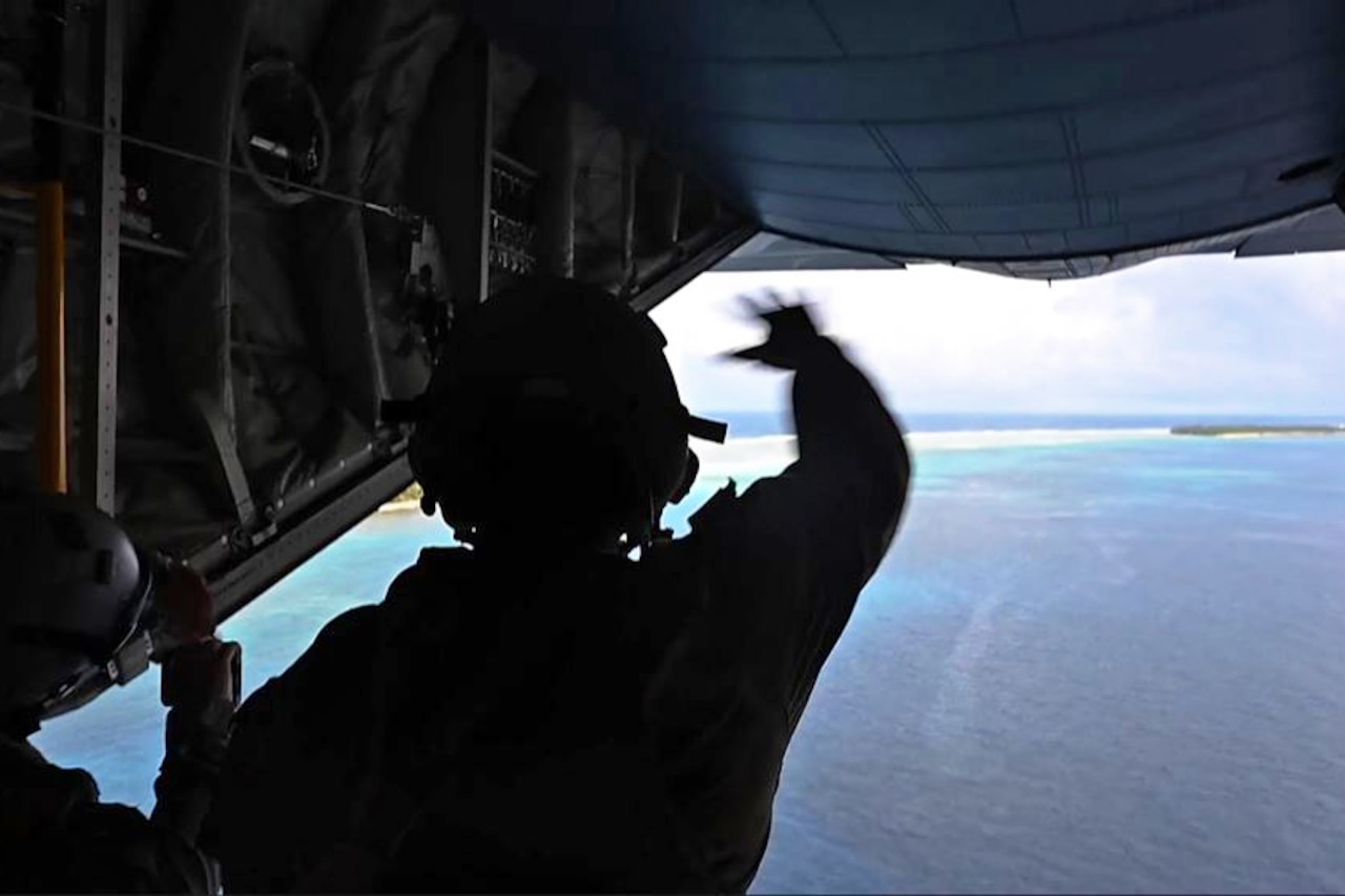 A service member waves toward an island from the back of an aircraft.