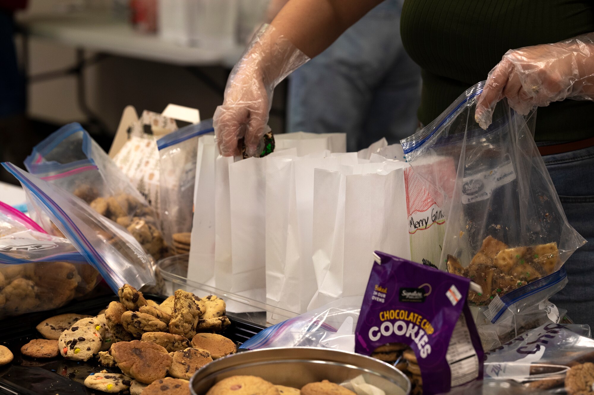 A pair of gloved hands are shown distributing cookies into white paper bags.