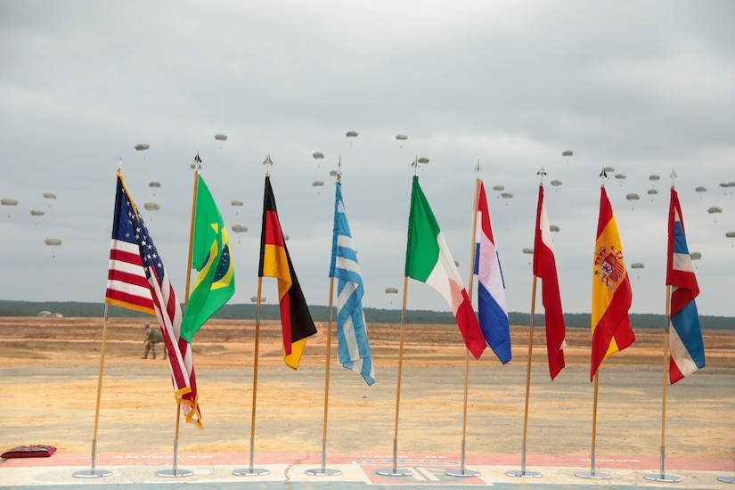 The flags of allied partner are proudly displayed on Sicily Drop Zone, Fort Bragg, NC, as paratroopers from the U.S. Civil Affairs Command (Airborne) alongside local Fort Bragg and partner nations' airborne Soldiers, gently drift from the sky during the the Randy Older Memorial Operation Toy drop 2.0, a combined airborne training event held from 4-12 DEC 2022 to increase joint airborne proficiency and enhance community relations.