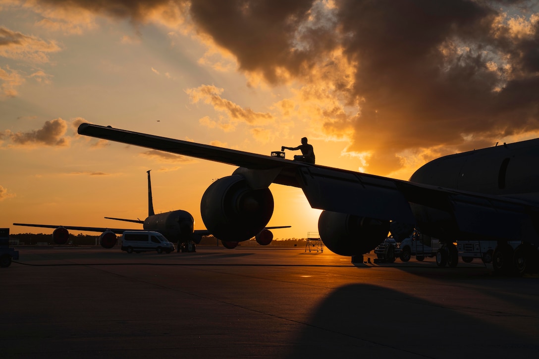 An airman works on the wing of an aircraft under a sunlit sky.