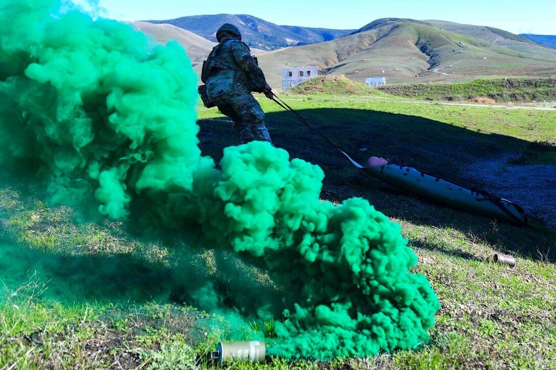 A soldier drags a dummy on a stretcher as green smoke rises from a can.