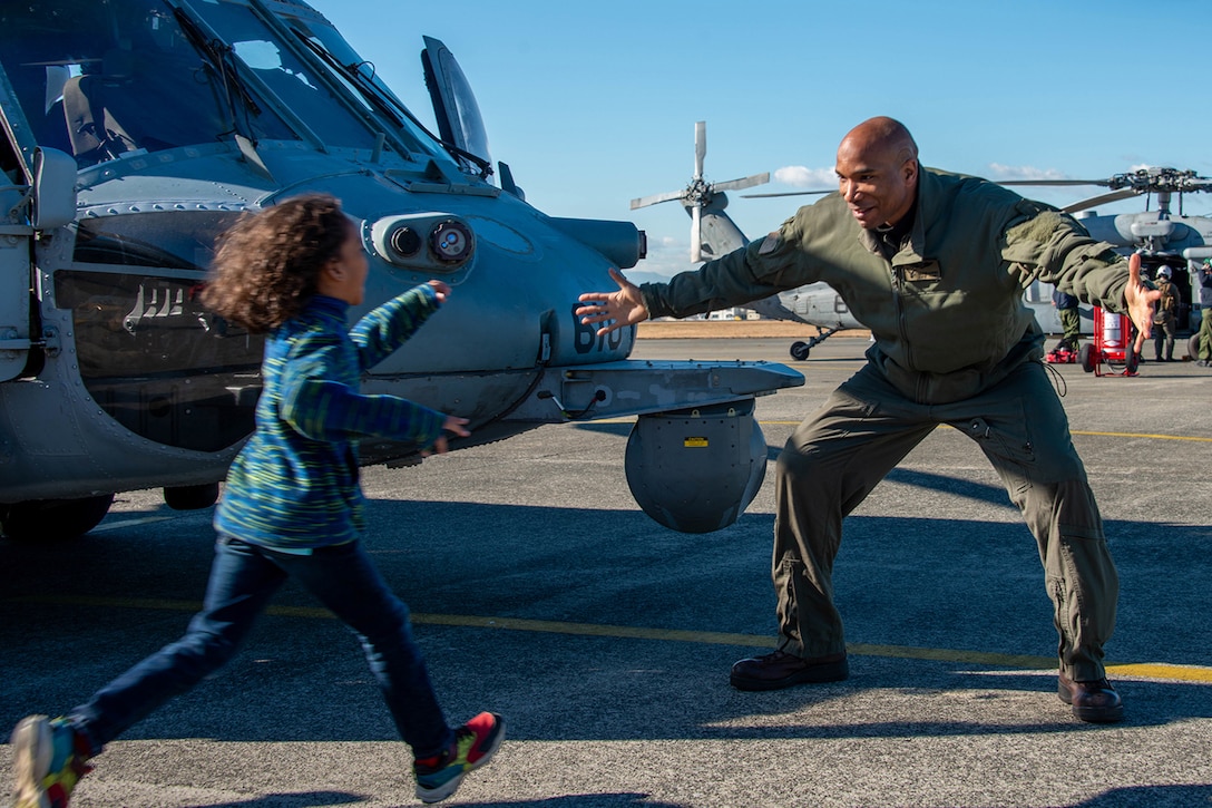 Man greets child with open arms while standing in front of military aircraft.