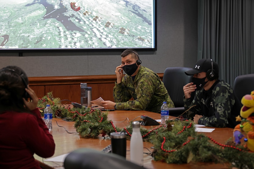 Three people sit at a conference table with Christmas decorations while monitoring a large screen on the wall.