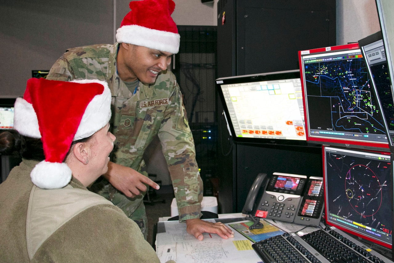 Two service members wearing Santa hats, one standing, converse while looking at monitors displaying radar graphics.