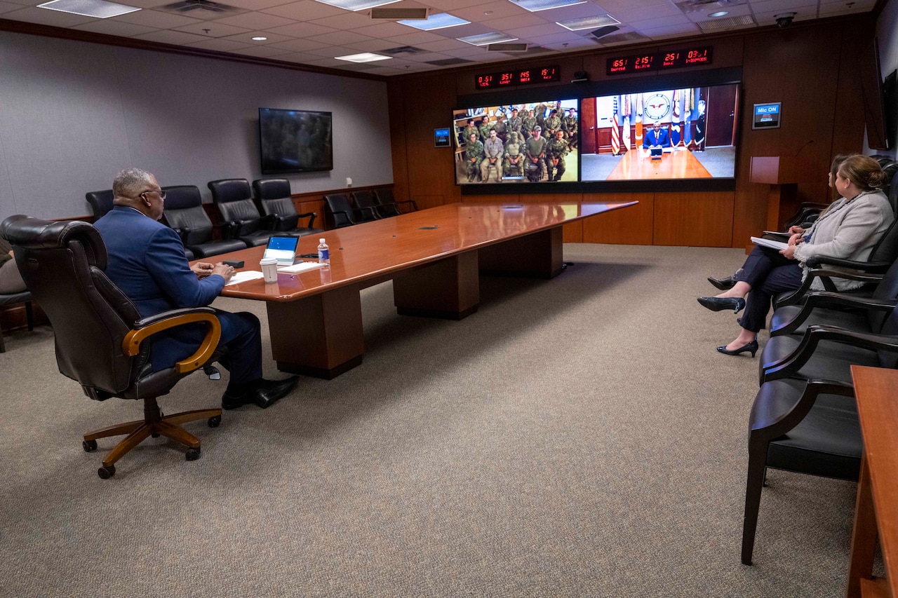 Secretary of Defense Lloyd J. Austin III speaks with airmen on a monitor from a large table in conference room with two other people seated on the sideline.