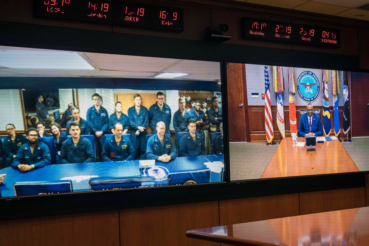 Secretary of Defense Lloyd J. Austin III is seen with sailors in a large split screen monitor.