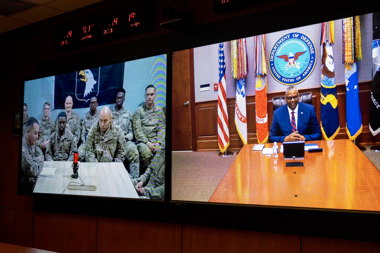 Secretary of Defense Lloyd J. Austin III smiles while looking at a laptop on a conference table with flags in the background.