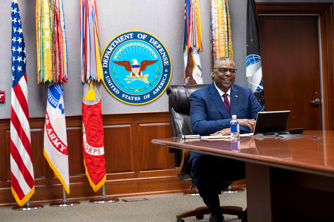 Secretary of Defense Lloyd J. Austin III smiles while looking at a laptop on a conference table with flags in the background.