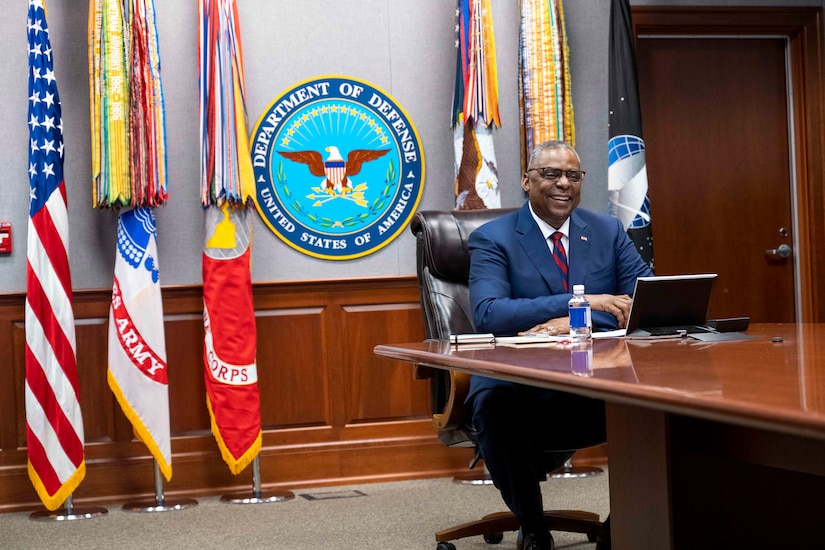 Secretary of Defense Lloyd J. Austin III smiles while looking at a laptop on a conference table with flags in the background.