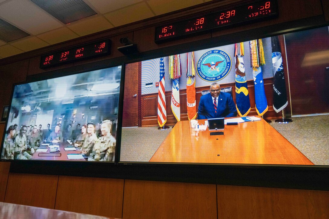 Secretary of Defense Lloyd J. Austin III is seen on a large monitor in split screen with a group of Marines.