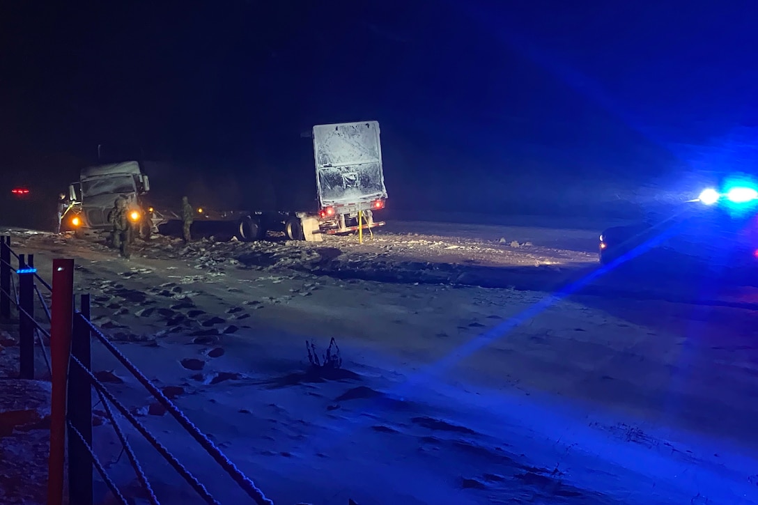 Guardsmen assist the driver of a truck on the side of a snowy road in the dark with blue light from an emergency response vehicle.