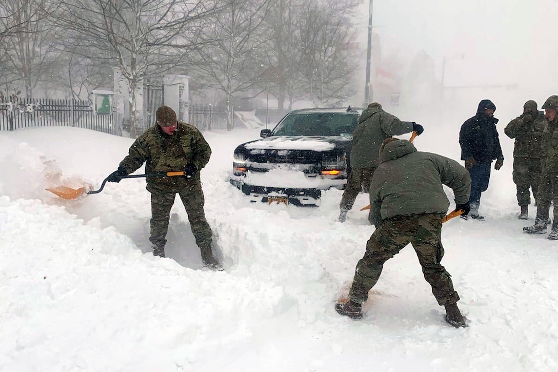 National Guardsmen shovel snow on a roadway in front of a civilian truck.