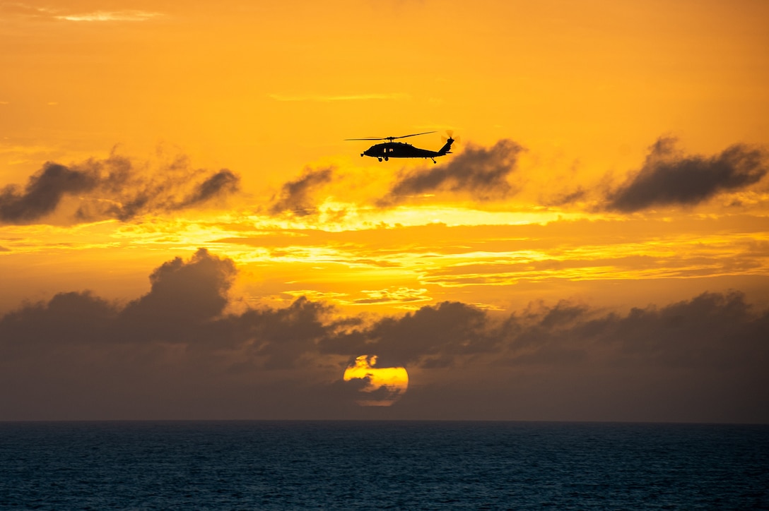 A military helicopter flies above an ocean with an orange sky in the background.