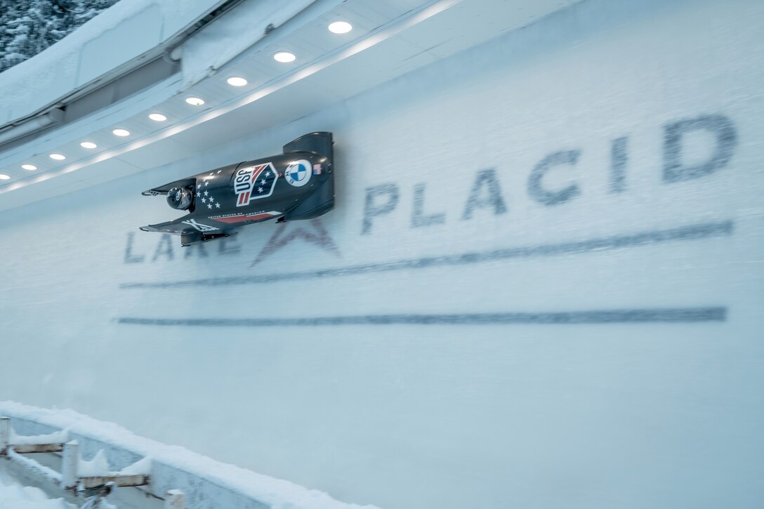 A bobsled navigates a curve on an ice track with "LAKE PLACID" written on the ice.