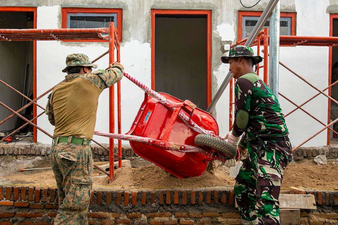 A Marine and an Indonesian service member dump dirt from a wheelbarrow at a construction site.