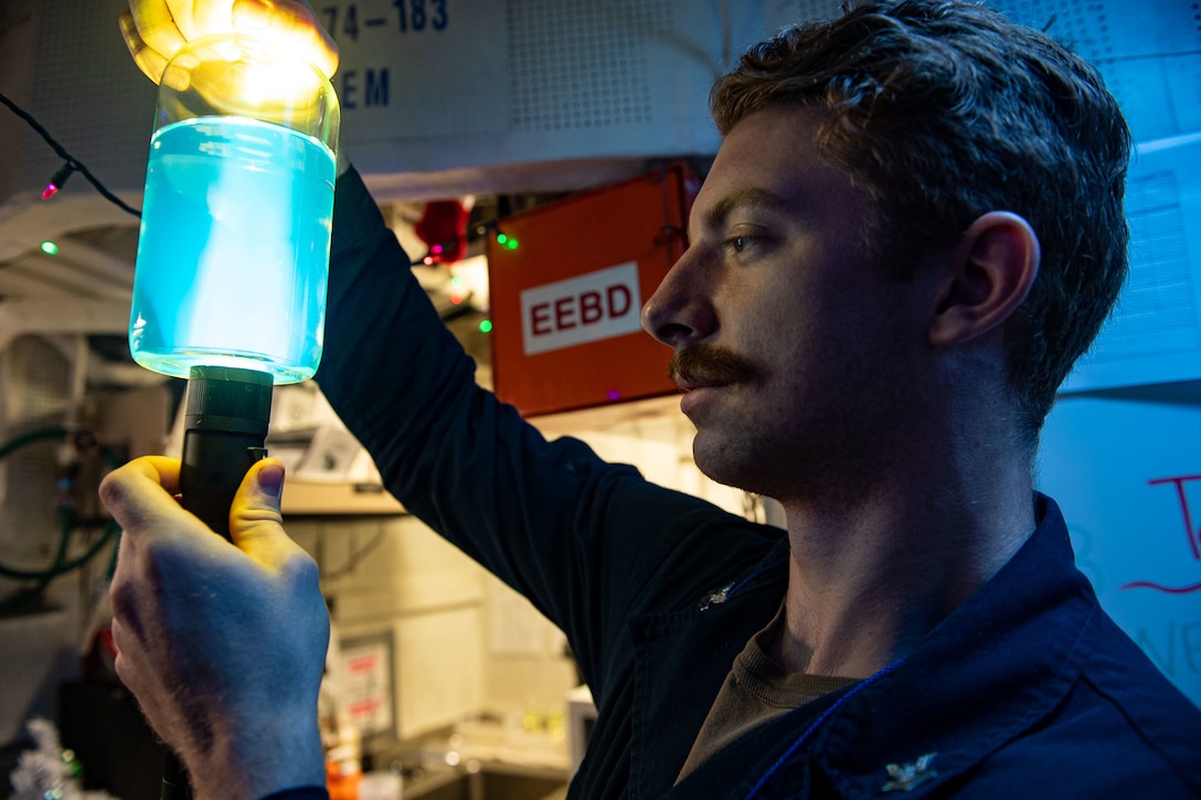 A sailor is shown close-up holding a fuel service tank sample.